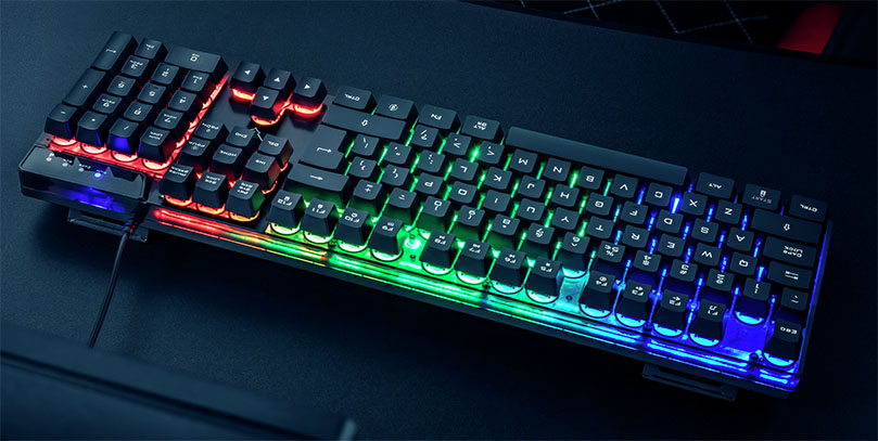 La imagen muestra un Teclado Trust GXT 835 Azor retroiluminado con luces LED multicolores sobre una mesa oscura.