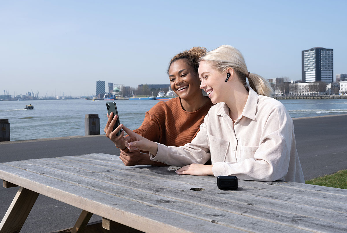 Dos personas sentadas en una mesa de madera al aire libre, con un teléfono móvil en las manos y mirando la pantalla juntos. Una de ellas está utilizando unos Auriculares inalámbricos Bluetooth Trust Primo Touch color negro. Al fondo se observa un cuerpo de agua y edificios urbanos bajo un cielo despejado.