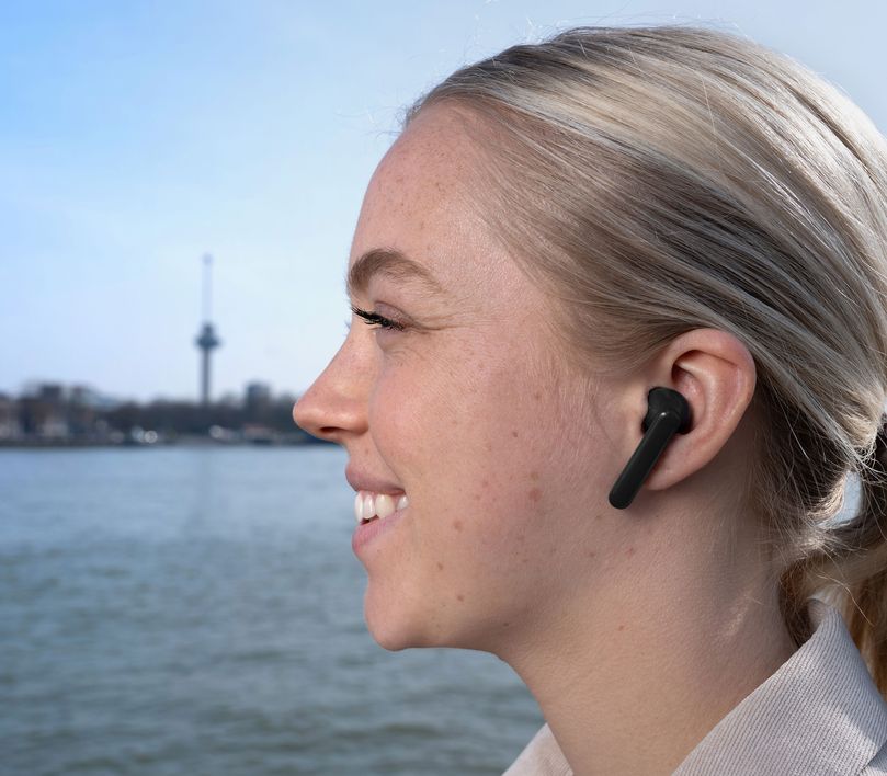 Una persona con cabello rubio recogido usa unos Auriculares inalámbricos Bluetooth Trust Primo Touch color negro. El fondo está desenfocado y muestra una torre de observación cerca de un cuerpo de agua.