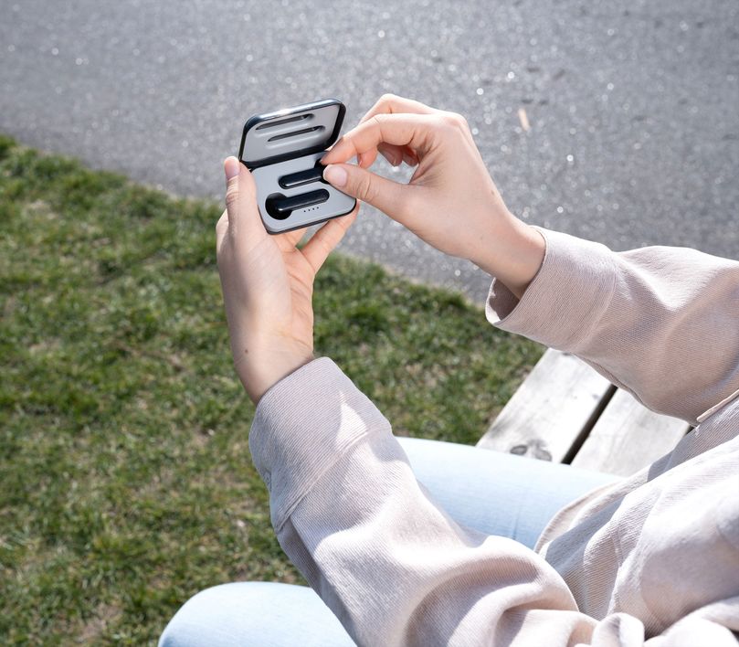 Una persona está al aire libre sosteniendo el estuche abierto de los Auriculares inalámbricos Bluetooth Trust Primo Touch color negro. La persona lleva una sudadera de color claro, y se ve césped en el fondo desenfocado.