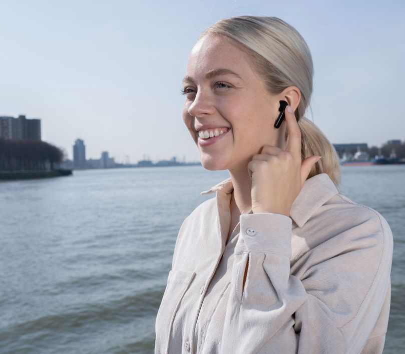 Persona con Auriculares inalámbricos Bluetooth Trust Primo Touch color negro tocando su oreja, con un río y edificios desenfocados en el fondo