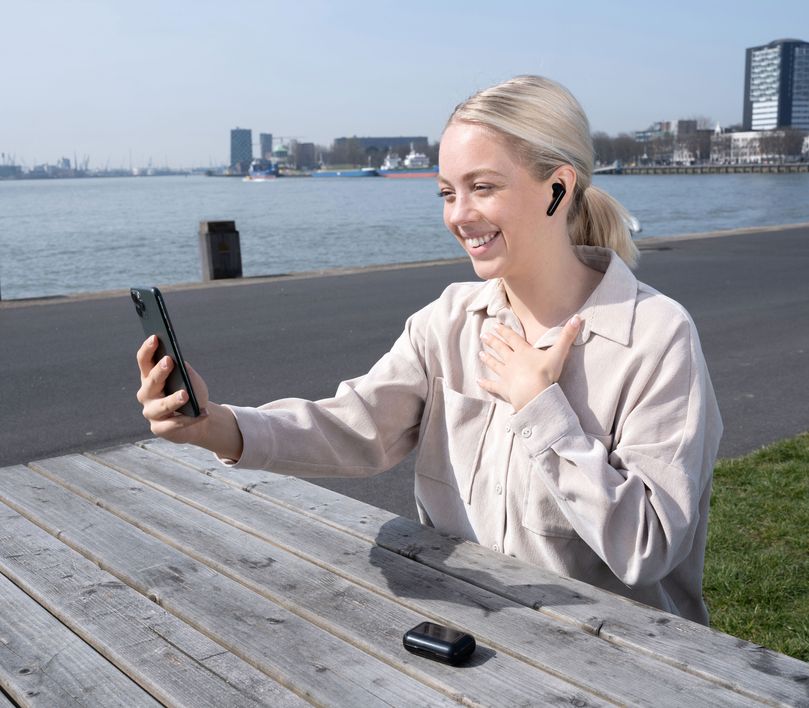 Una persona sentada en un banco al aire libre, con unos Auriculares inalámbricos Bluetooth Trust Primo Touch color negro, utilizando un teléfono inteligente. Al fondo, se ve un paisaje urbano con edificios y agua.