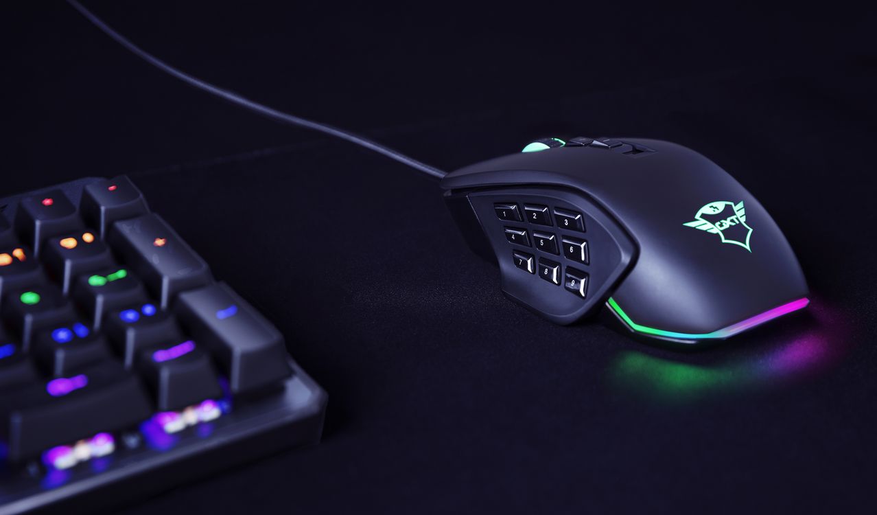 Claro, puedo ayudarte con eso. La imagen muestra un Mouse para gaming personalizable Trust GXT 970 Morfix con múltiples botones y luces LED de colores. Está junto a un teclado mecánico parcialmente iluminado sobre una superficie oscura.