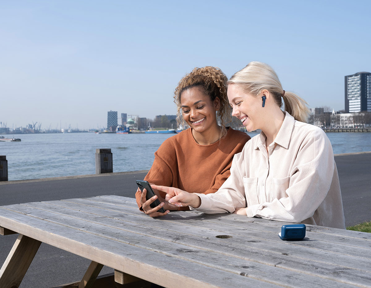 Dos personas sentadas en una mesa de madera al aire libre, con un teléfono móvil en las manos y mirando la pantalla juntos. Una de ellas está utilizando unos Auriculares inalámbricos Bluetooth Trust Primo Touch color azul. Al fondo se observa un cuerpo de agua y edificios urbanos bajo un cielo despejado.