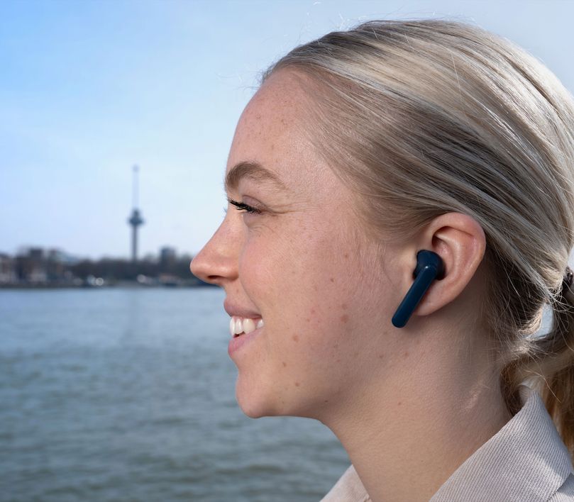 Una persona con cabello rubio recogido usa unos Auriculares inalámbricos Bluetooth Trust Primo Touch color azul. El fondo está desenfocado y muestra una torre de observación cerca de un cuerpo de agua.