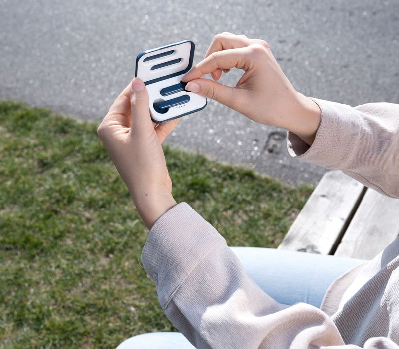 Una persona está al aire libre sosteniendo el estuche abierto de los Auriculares inalámbricos Bluetooth Trust Primo Touch color azul. La persona lleva una sudadera de color claro, y se ve césped en el fondo desenfocado.