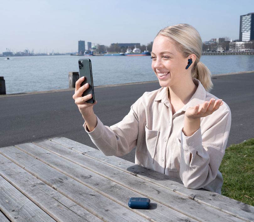 Una persona sentada en un banco al aire libre, con unos Auriculares inalámbricos Bluetooth Trust Primo Touch color azul, utilizando un teléfono inteligente. Al fondo, se ve un paisaje urbano con edificios y agua.