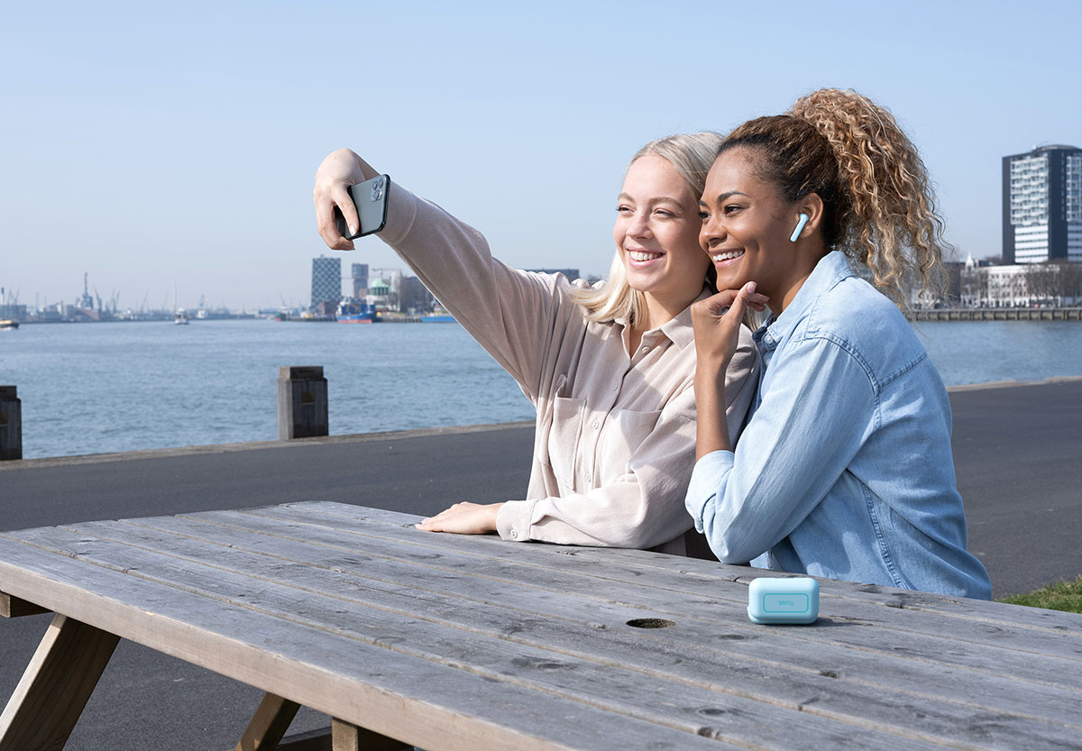 Dos personas sentadas en una mesa de madera al aire libre, con un teléfono móvil en las manos y mirando la pantalla juntos. Una de ellas está utilizando unos Auriculares inalámbricos Bluetooth Trust Primo Touch color turquesa. Al fondo se observa un cuerpo de agua y edificios urbanos bajo un cielo despejado.