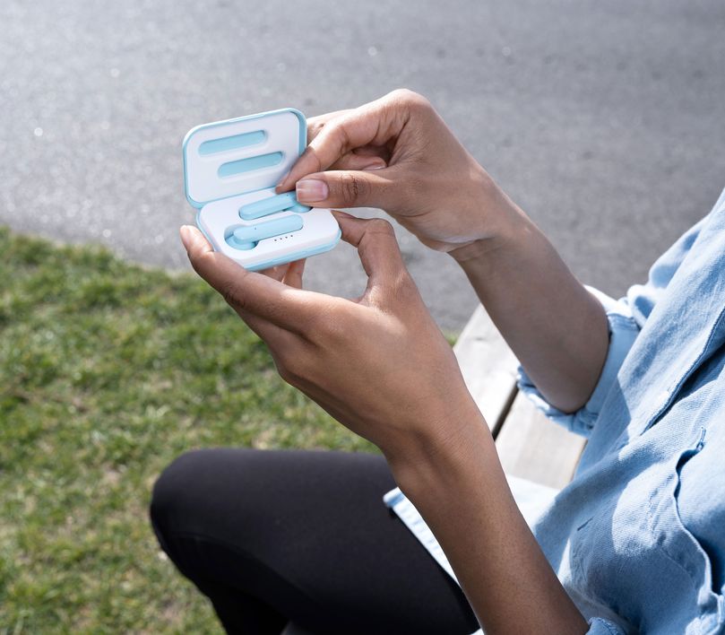 Una persona está al aire libre sosteniendo el estuche abierto de los Auriculares inalámbricos Bluetooth Trust Primo Touch color turquesa. La persona lleva una sudadera de color claro, y se ve césped en el fondo desenfocado.