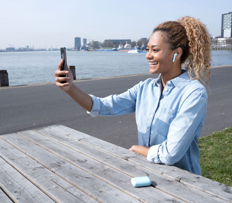 Una persona sentada en un banco al aire libre, con unos Auriculares inalámbricos Bluetooth Trust Primo Touch color turquesa, utilizando un teléfono inteligente. Al fondo, se ve un paisaje urbano con edificios y agua.