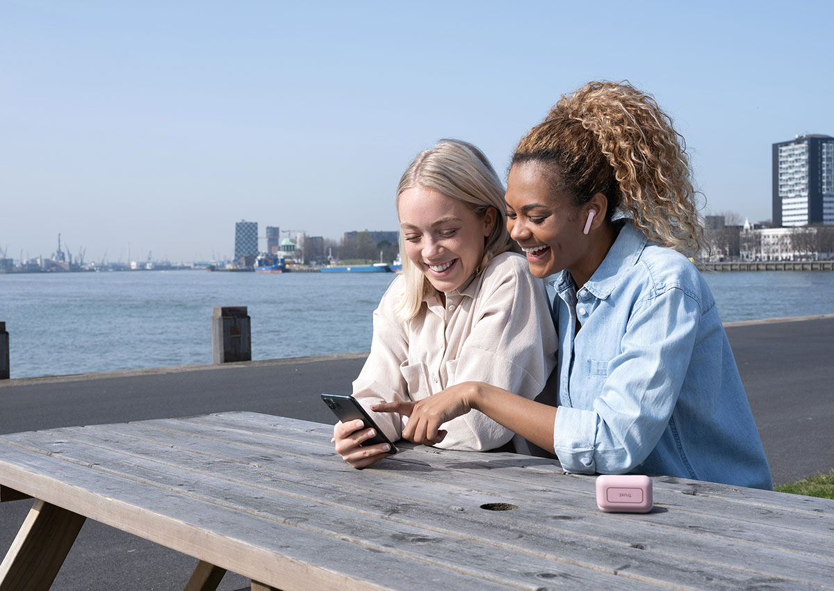 Dos personas sentadas en una mesa de madera al aire libre, con un teléfono móvil en las manos y mirando la pantalla juntos. Una de ellas está utilizando unos Auriculares inalámbricos Bluetooth Trust Primo Touch color rosado. Al fondo se observa un cuerpo de agua y edificios urbanos bajo un cielo despejado.