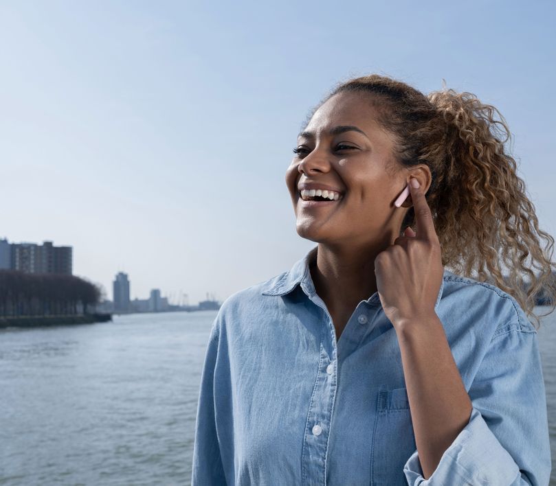 Persona con Auriculares inalámbricos Bluetooth Trust Primo Touch color rosado tocando su oreja, con un río y edificios desenfocados en el fondo