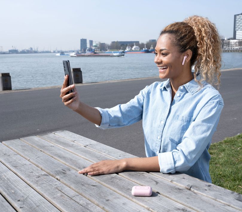 Una persona sentada en un banco al aire libre, con unos Auriculares inalámbricos Bluetooth Trust Primo Touch color rosado, utilizando un teléfono inteligente. Al fondo, se ve un paisaje urbano con edificios y agua.