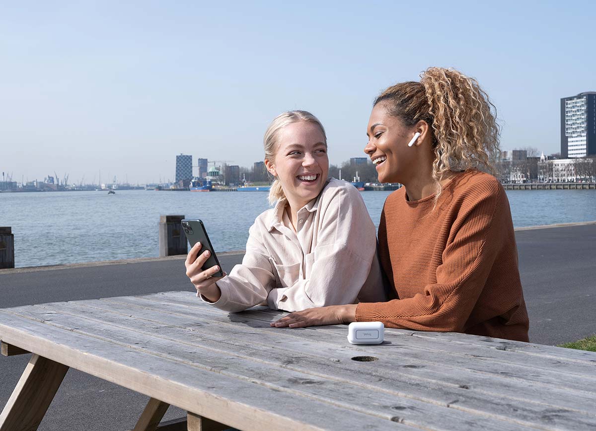 Dos personas sentadas en una mesa de madera al aire libre, con un teléfono móvil en las manos y mirando la pantalla juntos. Una de ellas está utilizando unos Auriculares inalámbricos Bluetooth Trust Primo Touch color blanco. Al fondo se observa un cuerpo de agua y edificios urbanos bajo un cielo despejado.