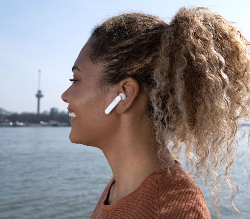 Una persona con cabello recogido usa unos Auriculares inalámbricos Bluetooth Trust Primo Touch color blanco. El fondo está desenfocado y muestra una torre de observación cerca de un cuerpo de agua.