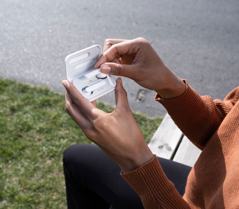 Una persona está al aire libre sosteniendo el estuche abierto de los Auriculares inalámbricos Bluetooth Trust Primo Touch color blanco. La persona lleva una sudadera de color claro, y se ve césped en el fondo desenfocado.