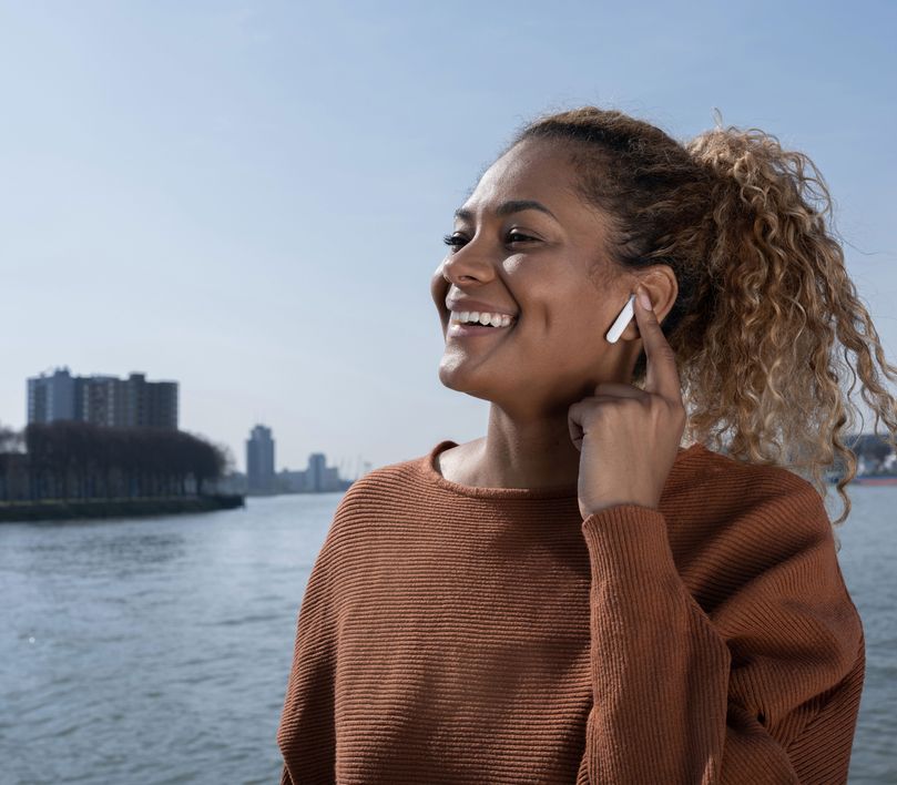 Persona con Auriculares inalámbricos Bluetooth Trust Primo Touch color blanco tocando su oreja, con un río y edificios desenfocados en el fondo