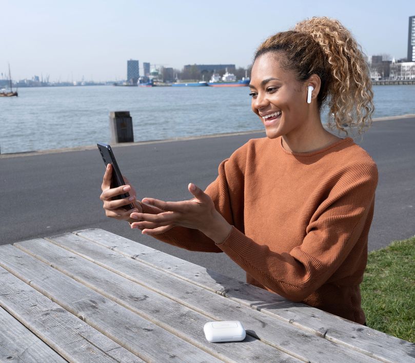 Una persona sentada en un banco al aire libre, con unos Auriculares inalámbricos Bluetooth Trust Primo Touch color blanco, utilizando un teléfono inteligente. Al fondo, se ve un paisaje urbano con edificios y agua.