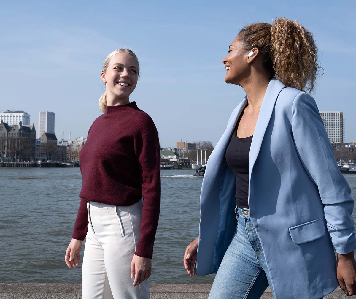 Dos personas caminando frente a un río con edificios en el fondo. Una de ellas tiene puestos unos Auriculares inalámbricos Bluetooth Trust Nika Compact color blanco