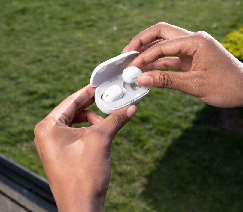 Una persona sosteniendo un estuche de carga abierto con Auriculares inalámbricos Bluetooth Trust Nika Compact color blanco dentro, en primer plano, con un fondo desenfocado de césped verde.