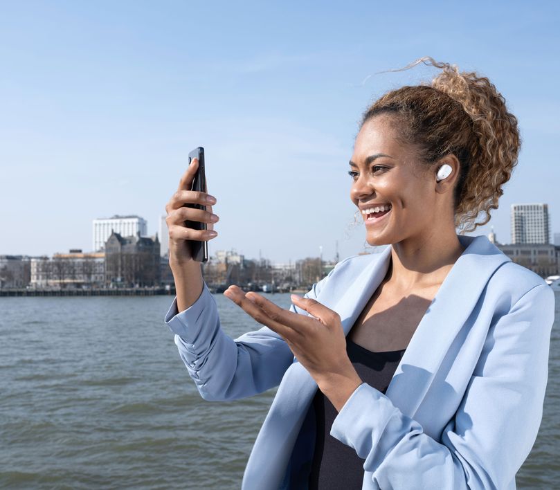 Una persona que lleva unos Auriculares inalámbricos Bluetooth Trust Nika Compact color blanco, sostiene un teléfono inteligente frente a un cuerpo de agua con edificios al fondo.
