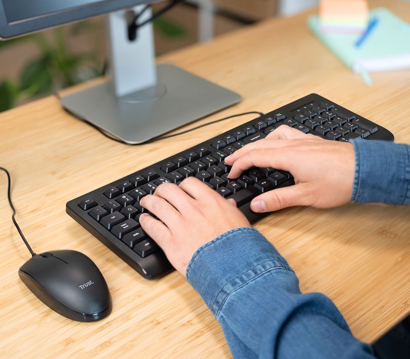 Manos utilizando un Combo de Teclado y Mouse Trust Primo en un escritorio, con una pantalla de computadora al fondo y útiles de oficina a la vista.