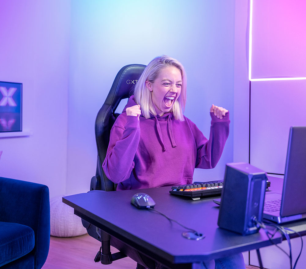 Persona con buzo sentada frente a un escritorio con una computadora, teclado, mouse y unos Parlantes 2.0 con iluminación RGB Trust GXT 609 Zoxa, levantando los brazos en un gesto de éxito o celebración. El ambiente tiene iluminación violeta y rosa.