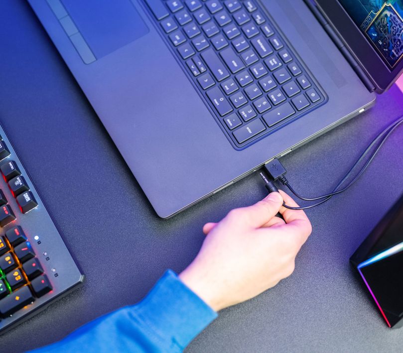 Una mano conectando el cable auxiliar de 3,5 mm de los Parlantes 2.0 con iluminación RGB Trust GXT 609 Zoxa a una laptop sobre un escritorio con iluminación tenue.
