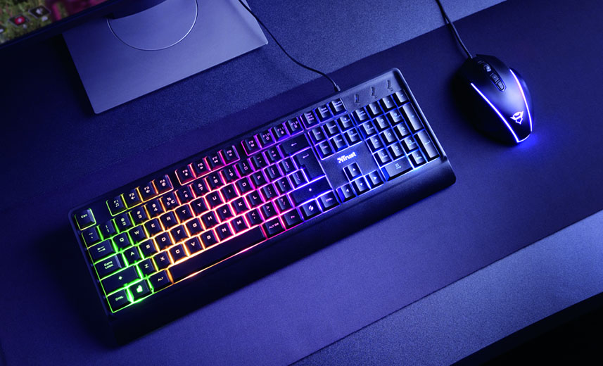 Teclado Trust Ziva y mouse de computadora con iluminación RGB en un escritorio. El teclado Ziva muestra una gama de colores que van del verde al rojo, pasando por el amarillo, el azul y el morado. El mouse también tiene iluminación LED, destacando con un borde azul brillante.