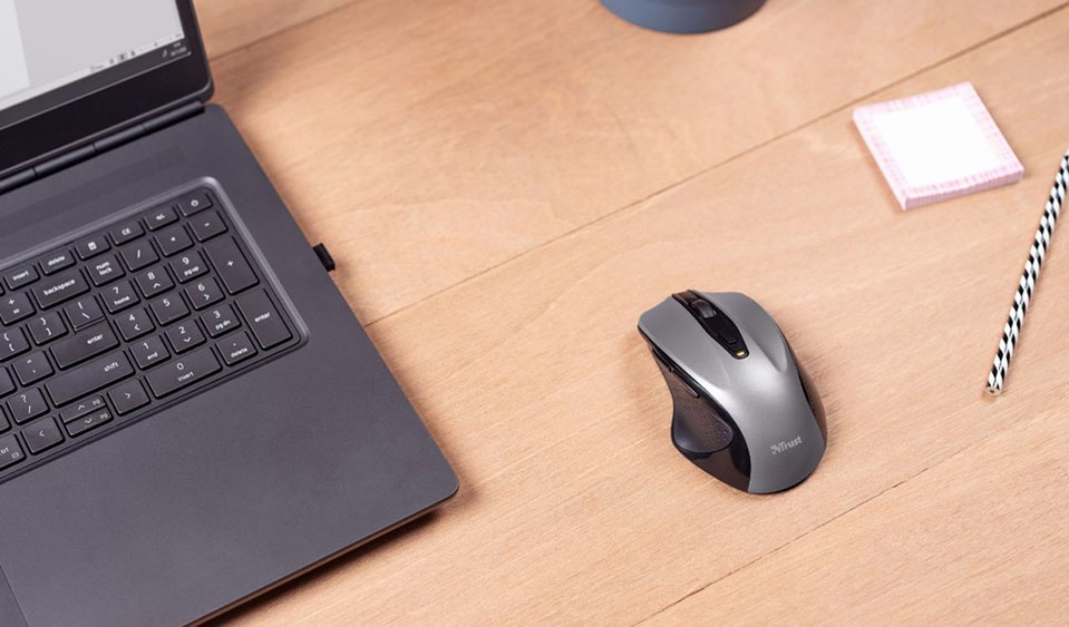 Una computadora portátil descansa sobre una mesa de madera. A su lado derecho, se encuentra un Mouse inalámbrico Trust Nito, un bloc de notas rosa y un lápiz con diseño de rayas en la esquina superior derecha.
