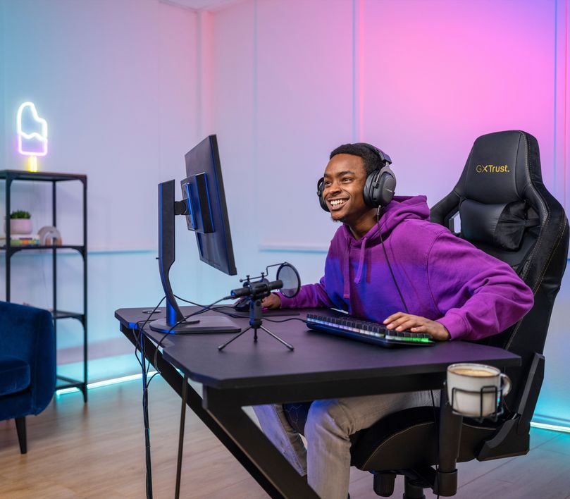 Una persona está sentada frente a una computadora con auriculares, usando un Micrófono USB para streaming Trust GXT 241 Velica, un teclado y un mouse. La habitación está iluminada con luces LED de colores. La silla es de color negro y tiene el logo de “Trust”. Al fondo se observa una planta y un neón en forma de helado.