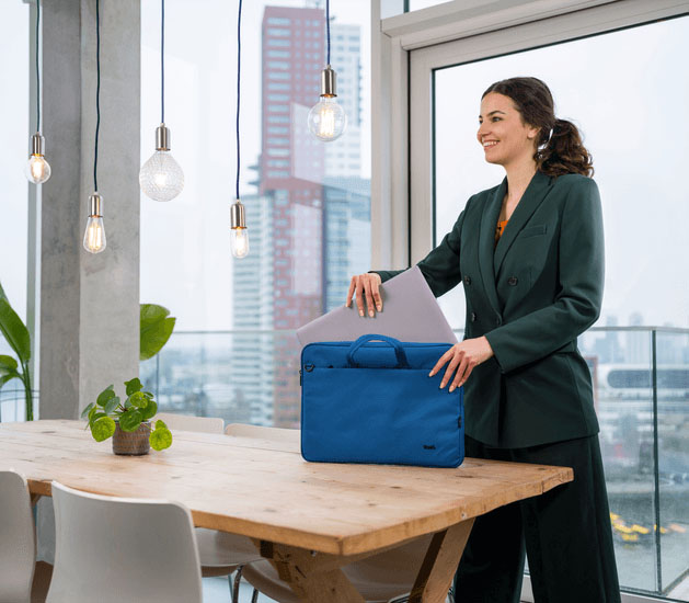 Una mujer joven y sonriente con un traje saca una laptop gris del maletín Trust Bologna Eco-friendly Slim azul de 16 pulgadas, que está sobre una mesa de madera rústica con grandes ventanales de fondo.