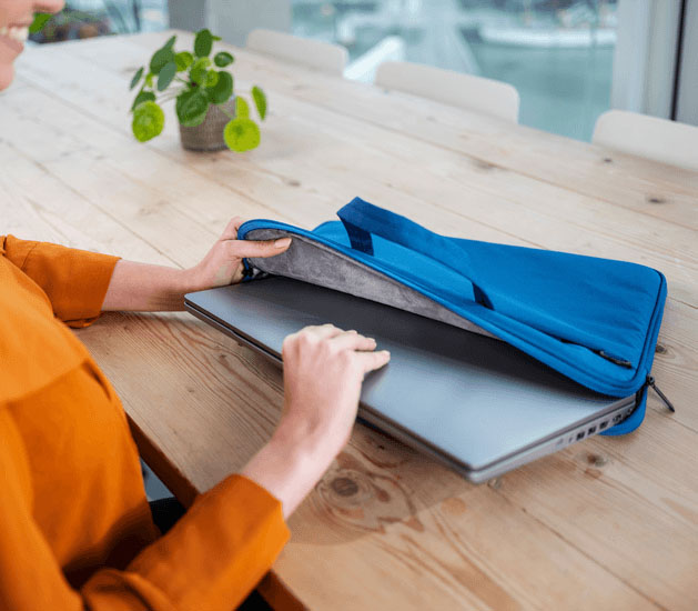 Una toma de cerca de las manos de una mujer insertando una laptop plateada en el compartimento principal acolchado del maletín Trust Bologna Eco-friendly Slim azul de 16 pulgadas, que está sobre una mesa de madera.