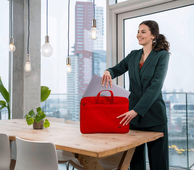 Una mujer joven y sonriente con un traje saca una laptop gris del maletín Trust Bologna Eco-friendly Slim rojo de 16 pulgadas, que está sobre una mesa de madera rústica con grandes ventanales de fondo.