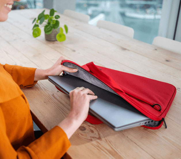 Una toma de cerca de las manos de una mujer insertando una laptop plateada en el compartimento principal acolchado del maletín Trust Bologna Eco-friendly Slim rojo de 16 pulgadas, que está sobre una mesa de madera.