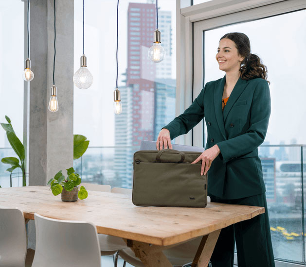 Una mujer joven y sonriente con un traje saca una laptop gris del maletín Trust Bologna Eco-friendly Slim verde de 16 pulgadas, que está sobre una mesa de madera rústica con grandes ventanales de fondo.