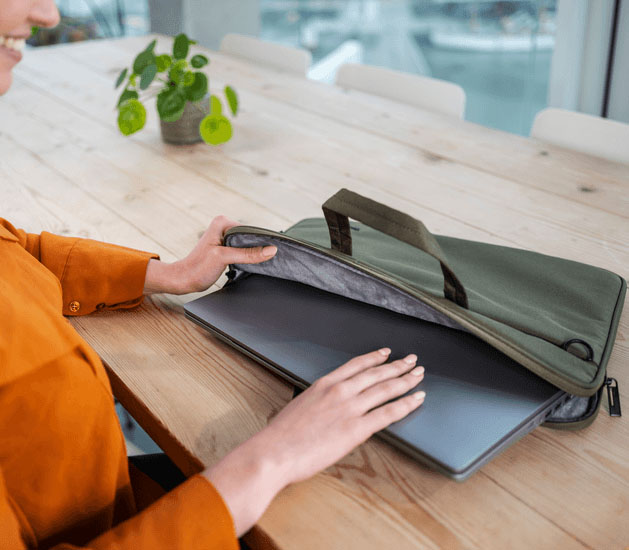 Una toma de cerca de las manos de una mujer insertando una laptop plateada en el compartimento principal acolchado del maletín Trust Bologna Eco-friendly Slim verde de 16 pulgadas, que está sobre una mesa de madera.