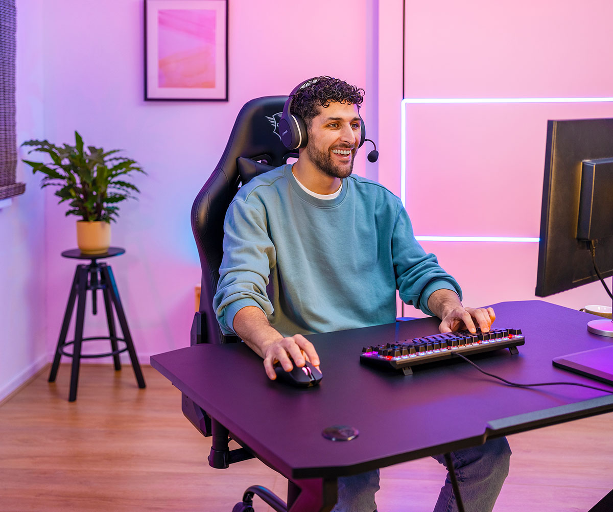 Persona sentada frente a un escritorio con una computadora, en una habitación iluminada con luces rosadas y azules. La persona utiliza un Mouse gaming inalámbrico recargable Trust GXT 980 Redex, con un teclado mecánico al frente. Hay una planta en la esquina izquierda y cuadros decorativos en la pared.