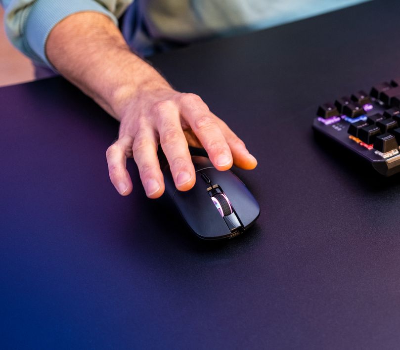 La imagen muestra una mano sobre un Mouse gaming inalámbrico recargable Trust GXT 980 Redex negro al lado de un teclado mecánico en un escritorio.