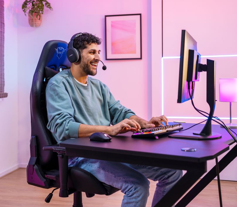 Persona sentada frente a un escritorio utilizando una computadora en una habitación iluminada con luces rosadas y moradas. La persona lleva puestos auriculares, y está tocando el teclado cerca de un Mouse gaming inalámbrico recargable Trust GXT 980 Redex. Hay cuadros decorativos en la pared detrás.