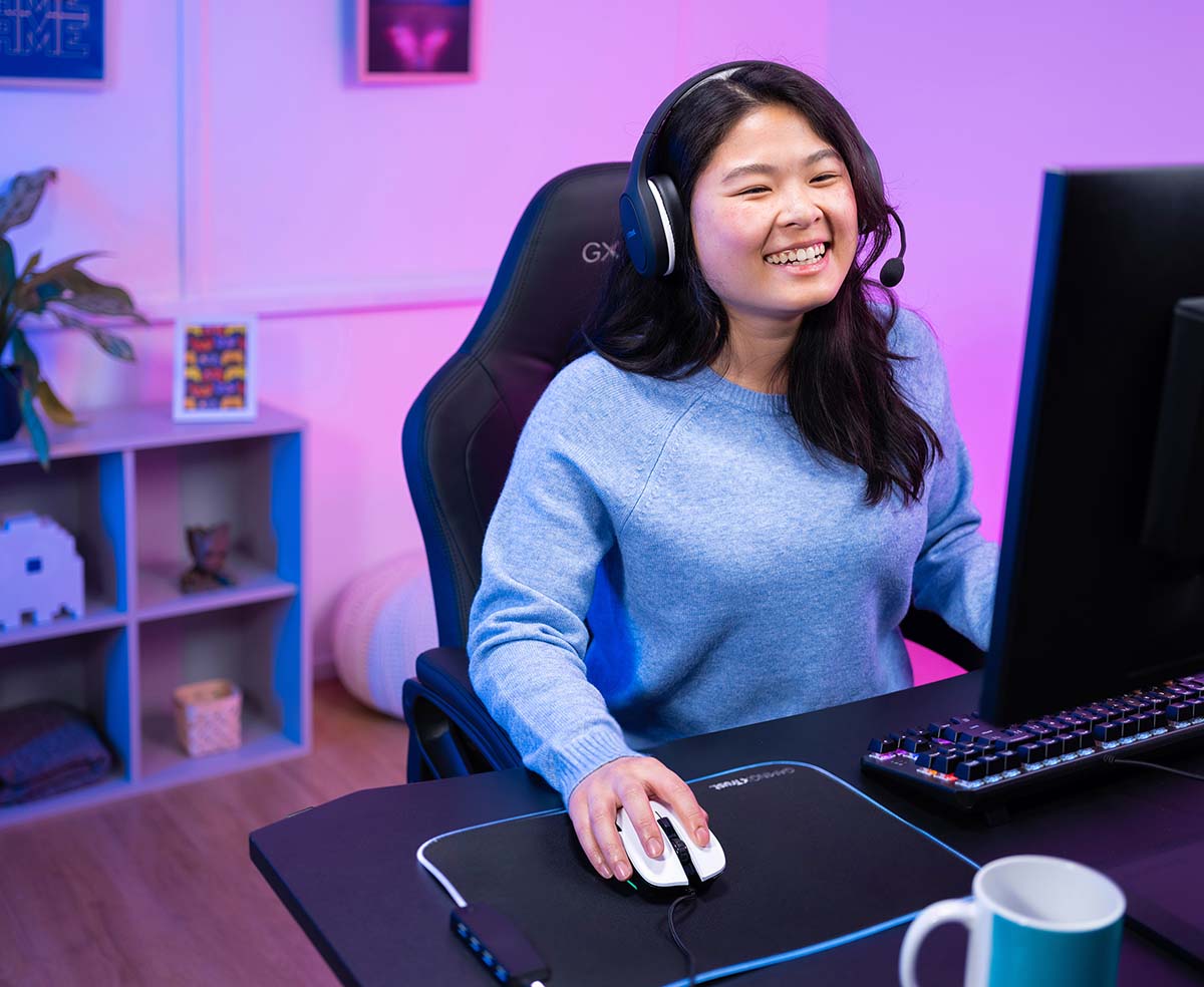 La imagen muestra a una persona sentada frente a una computadora. Tiene una mano en el teclado y la otra en un Mouse gaming Trust GXT 922W Ybar de color blanco. La iluminación alrededor es de tonos azules y violetas.
