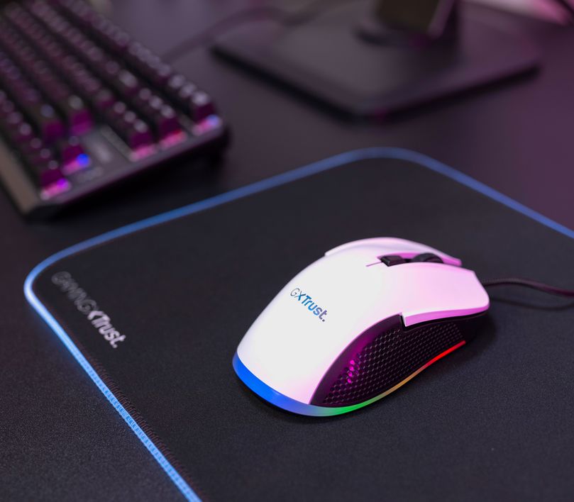 Mouse gaming Trust GXT 922W Ybar de color Blanco sobre un mousePad con iluminación LED multicolor, junto a un teclado parcialmente visible en el fondo, todo sobre un escritorio con una superficie oscura.