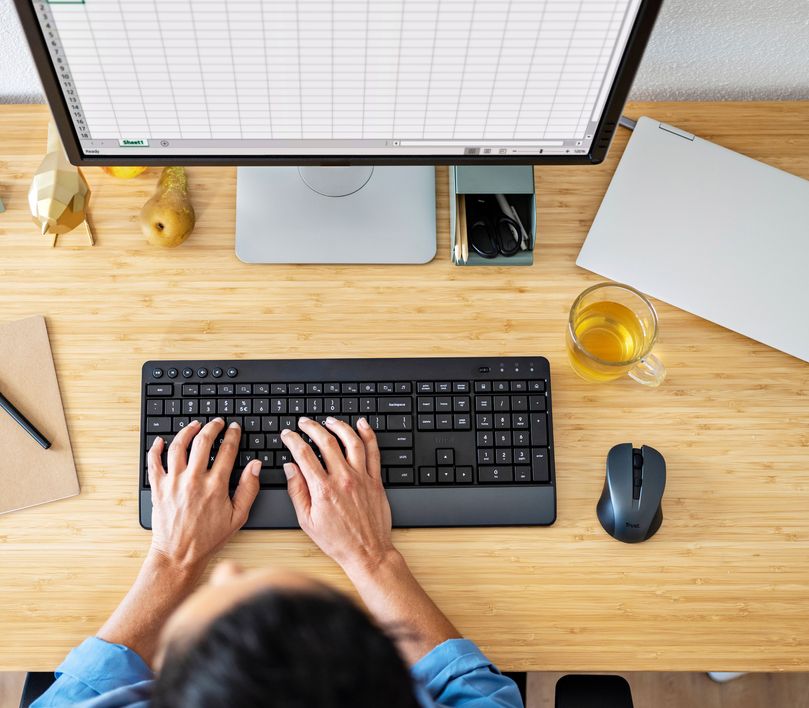 Vista aérea de una persona utilizando un Combo de Teclado y Mouse inalámbrico Trust Trezo Comfort en un escritorio con una computadora de escritorio, una taza de té, y otros objetos.
