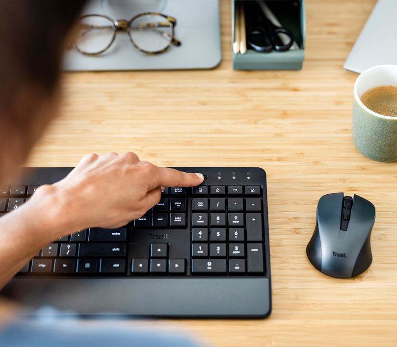 Persona trabajando en una computadora portátil con un Combo de Teclado y Mouse inalámbrico Trust Trezo Comfort, anteojos y una taza de café sobre el escritorio.