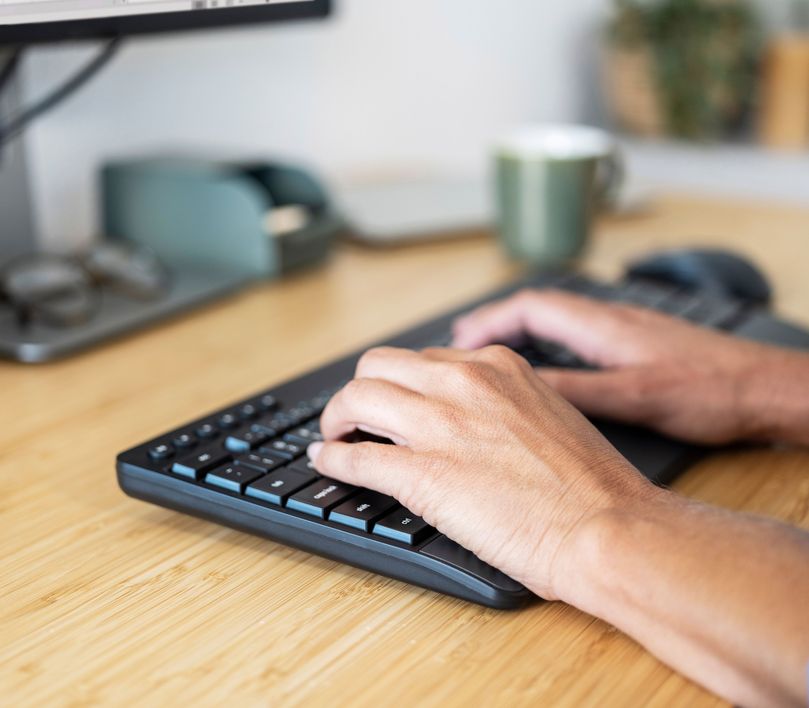 Manos sobre el teclado de un Combo de Teclado y Mouse inalámbrico Trust Trezo Comfort en una oficina con una computadora y una taza al fondo.