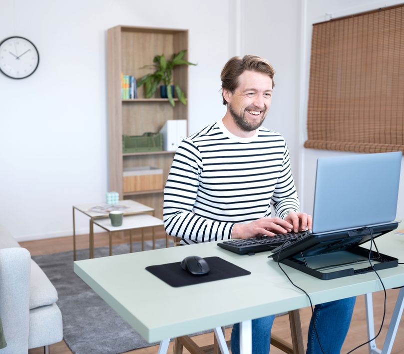 Persona sentada frente a una computadora portátil en una Base de refrigeración para Notebook Trust Exto sobre un escritorio, con un teclado y un mouse adicionales, en una habitación bien iluminada con muebles modernos y un reloj en la pared.