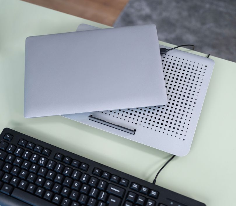 Una computadora portátil cerrada sobre una Base de refrigeración Trust Exto, ambas colocadas sobre un escritorio junto a un teclado.