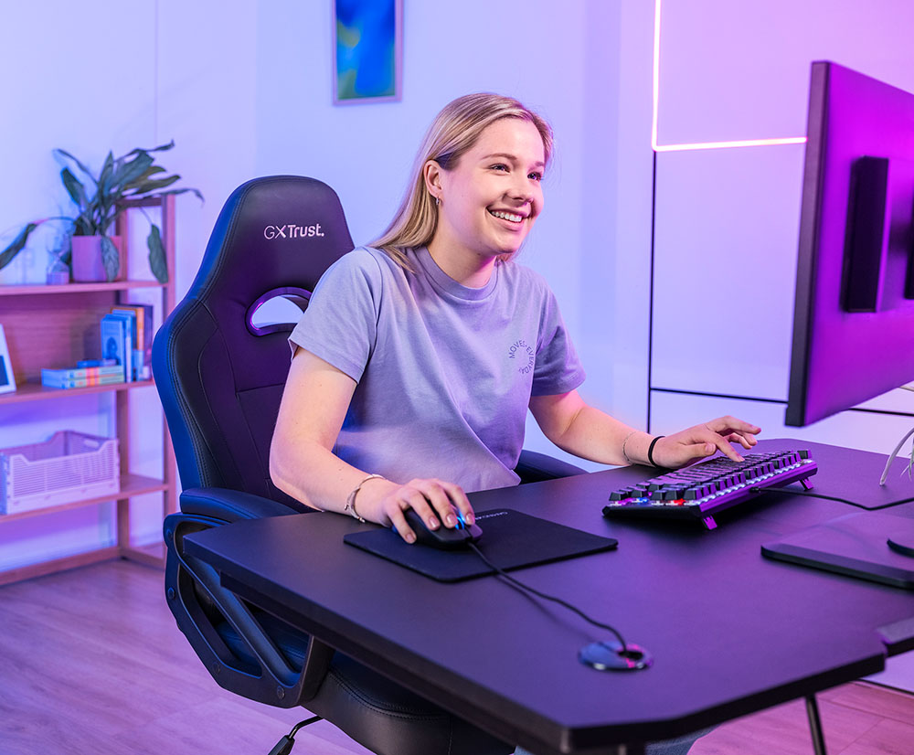 La imagen muestra a una persona sentada frente a un escritorio. Está utilizando una computadora con un Mouse gaming ligero Trust GXT 981 Redex y un teclado iluminado. La habitación está iluminada con luces violetas. La persona tiene cabello rubio y viste una remera celeste. Se encuentra en una silla gamer negra con el logo de GX Trust.