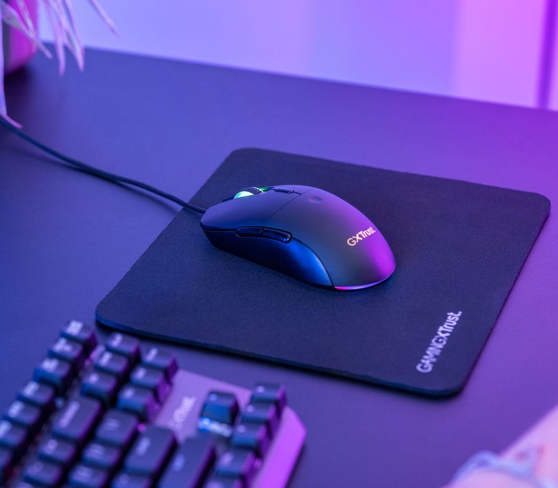 La imagen muestra un Mouse gaming ligero Trust GXT 981 Redex con iluminación RGB sobre un mousepad negro. Junto a él, se ve parcialmente un teclado mecánico. Todo está colocado en un escritorio iluminado con uan tenue luz violeta.