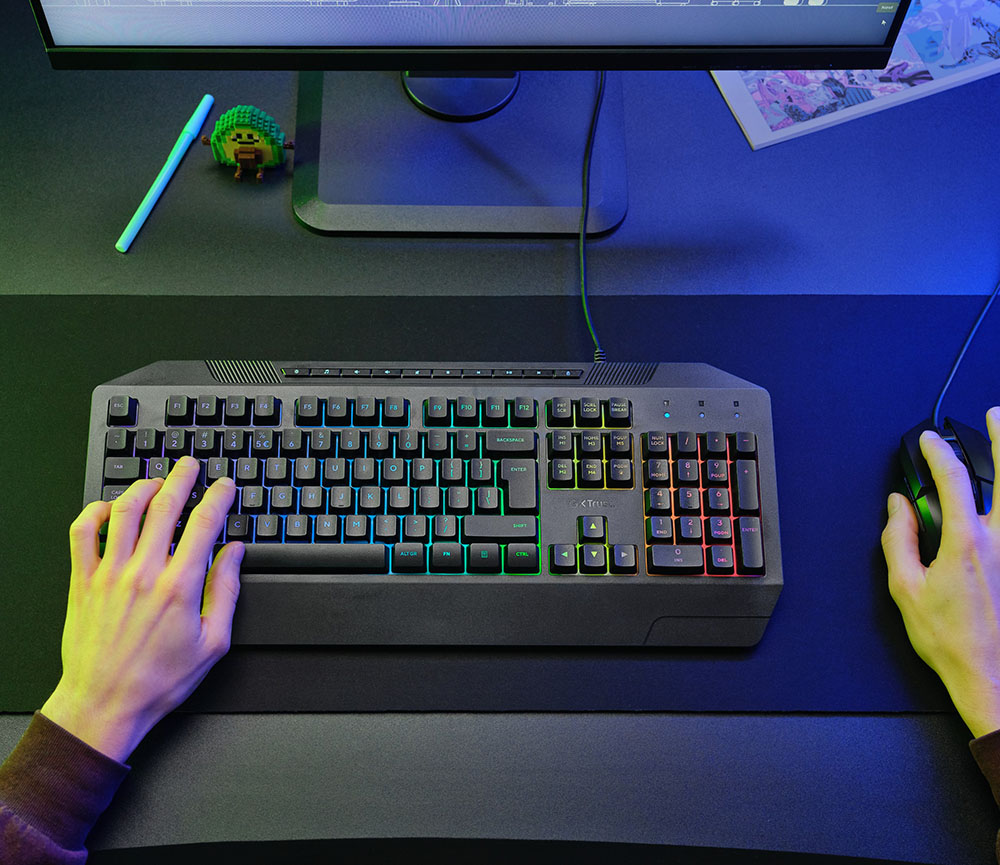 Teclado Gaming Iluminado Trust GXT 836 Evocx sobre un escritorio, manos utilizando el teclado y un mouse, monitor encendido al fondo y objetos decorativos alrededor.